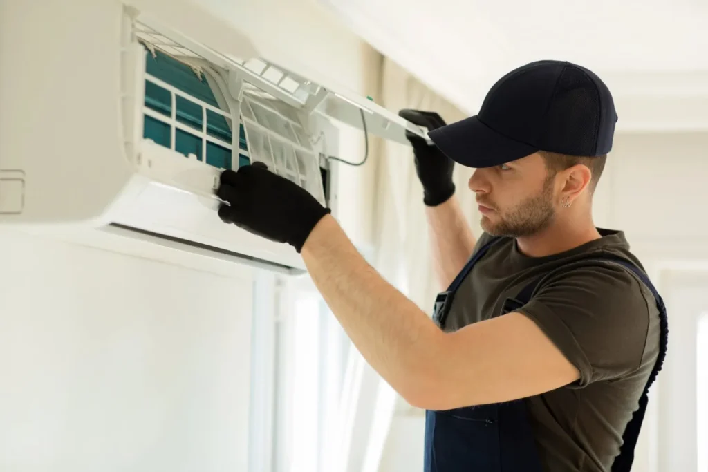 A service technician wearing a cap and black gloves is inspecting or working on the internal components of a split AC unit.