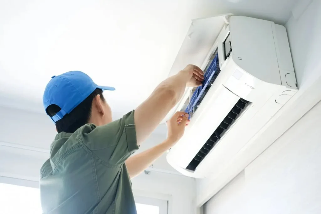 A technician in a blue cap is cleaning or repairing the air filter of a wall-mounted indoor AC unit.
